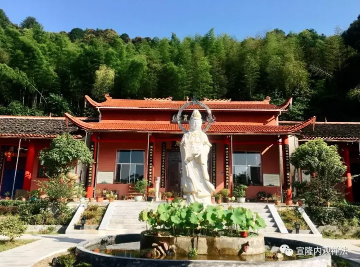 A red temple hall and pale statue beside a pond and wooded hillside.
