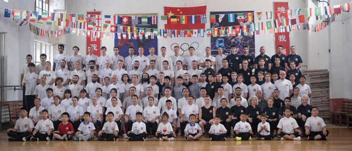 A large academy group poses beneath flags in an indoor hall.