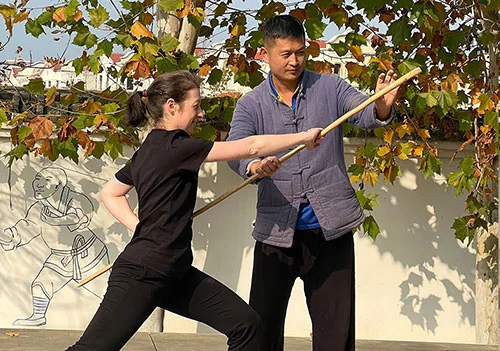 A teacher guides a student’s staff technique outdoors.