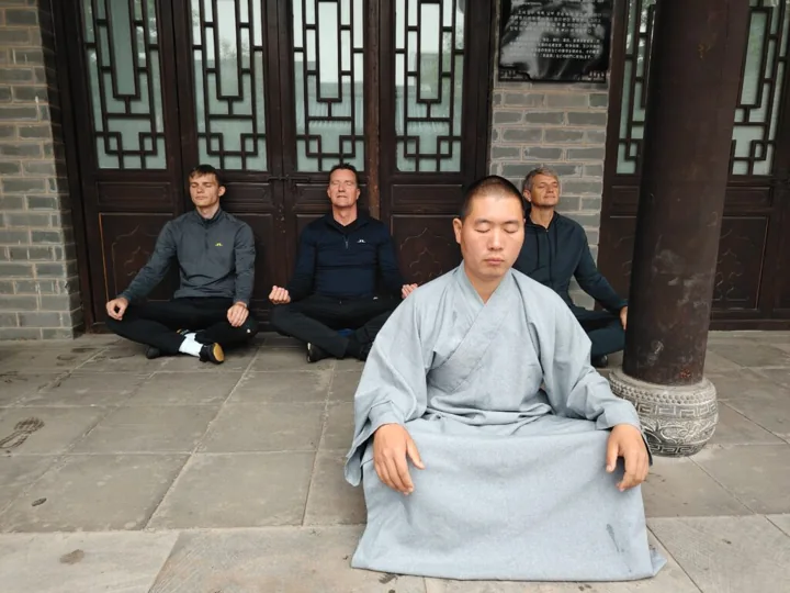 Students in grey robes meditate beside wooden columns.