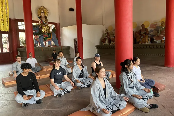 Students sit cross-legged inside a temple hall with red columns.