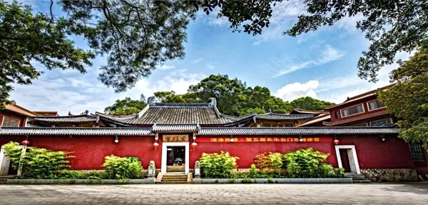 A long red temple wall and gate beneath leafy trees.