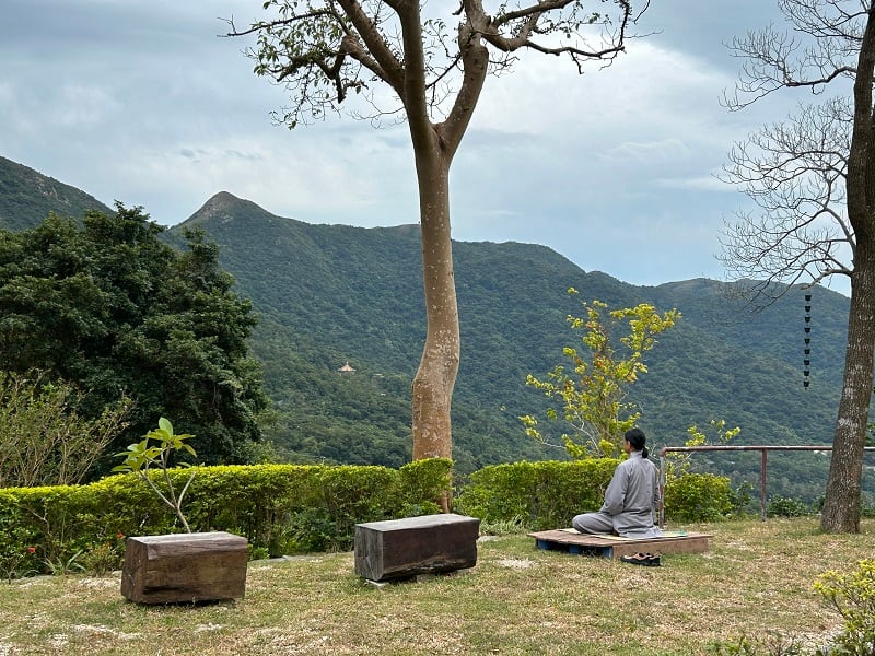 Ein Meditierender im Gewand sitzt auf einer Wiese vor den Gipfeln von Lantau.