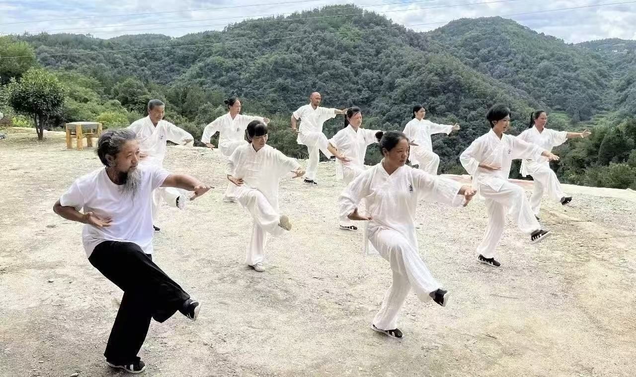 Adults in white practise tai chi on an open platform below wooded ridges.