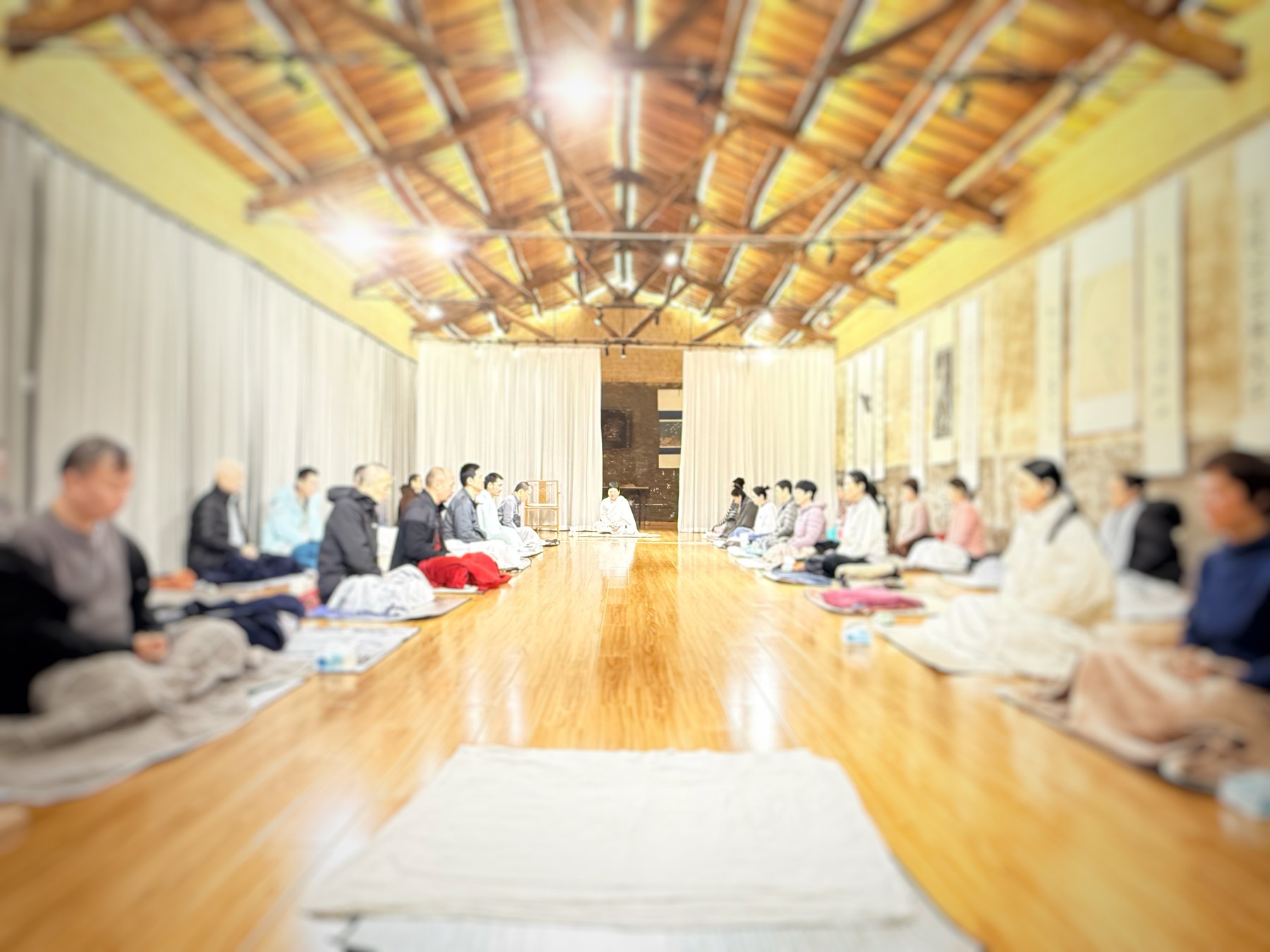 Participants sit in two rows inside a bright wooden-roofed hall.
