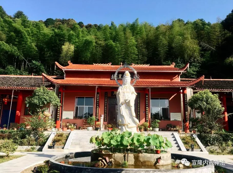 A red temple hall and pale statue beside a pond and wooded hillside.