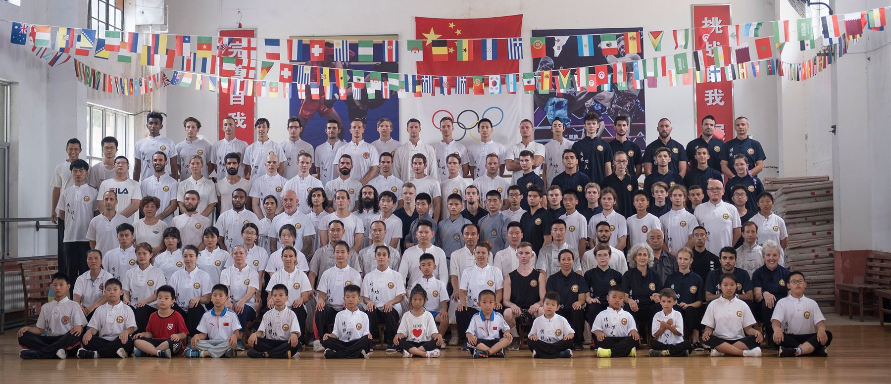 A large academy group poses beneath flags in an indoor hall.
