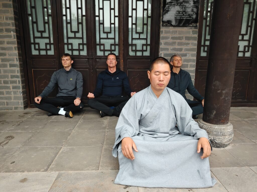 Studenti in vesti grigie meditano accanto a colonne di legno.