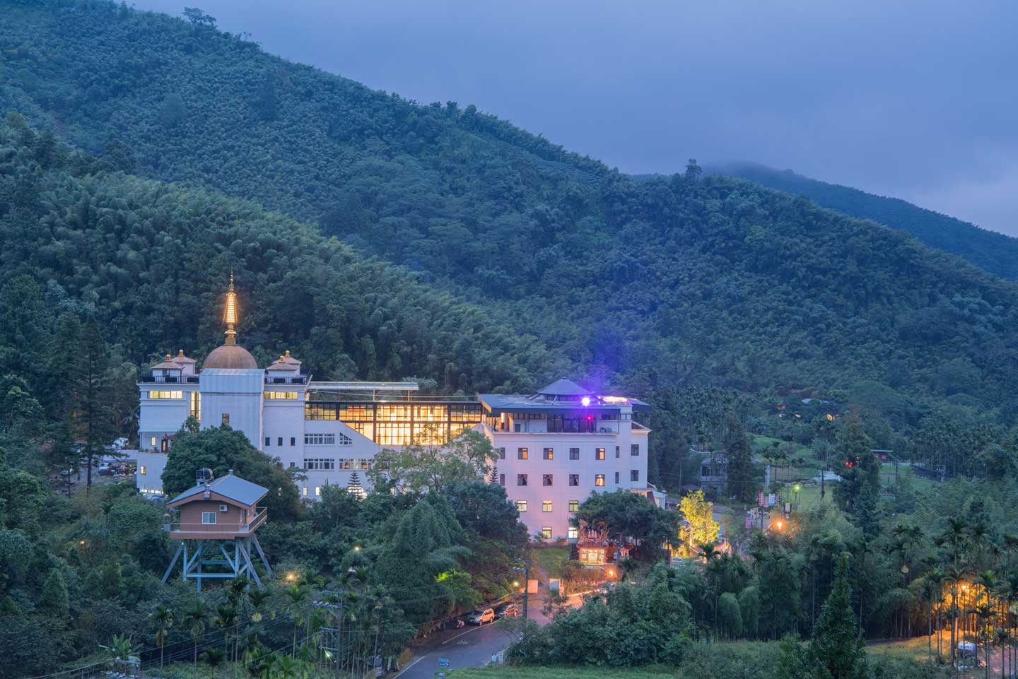 A monastery and golden stupa at dusk below misty forested hills.